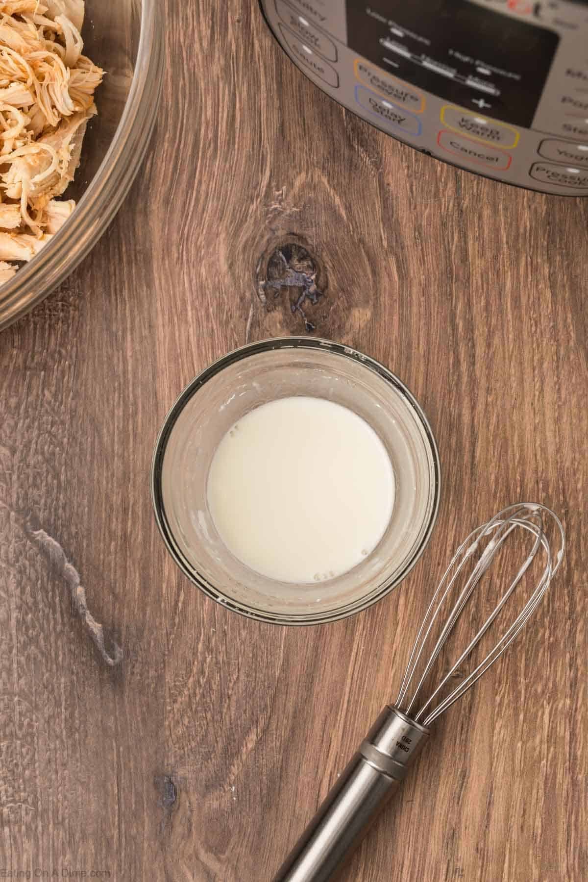 A small glass bowl of cornstarch slurry sits on a wooden surface beside a metal whisk, a bowl of shredded chicken, and part of an electric pressure cooker—perfect for making Instant Pot Buffalo Chicken Pasta.
