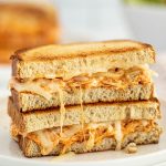 A close-up of a Buffalo Chicken Grilled Cheese sandwich with shredded chicken and melted cheese, cut in half and stacked on a white plate. The golden brown bread looks crispy with gooey cheese oozing out.