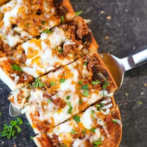 Sloppy Joe Stuffed French Bread features sliced French bread topped with melted cheese, ground beef, savory sauce, and chopped parsley, served on a dark surface with a spatula lifting a piece.