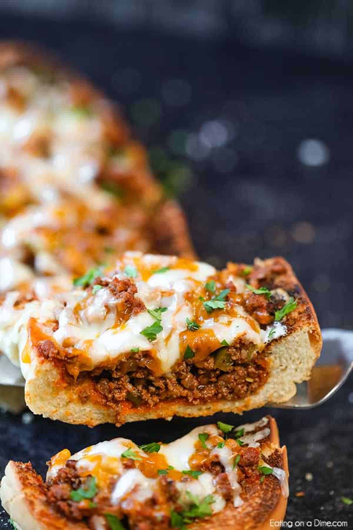 Sloppy joes stuffed french bread recipe has everything you love about sloppy joes in a cheese stuffed bread. This is the perfect party food or dinner idea. A close-up of cheesy sloppy joes stuffed French bread. The bread is loaded with a savory ground beef mixture, melted cheese, and garnished with chopped parsley. A slice is being lifted with a spatula, revealing the rich filling inside the crusty bread.