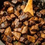 Close-up of Garlic Butter Steak Bites in a skillet, with a wooden spoon stirring the browned, juicy cubes of beef, seasoned with herbs and spices.