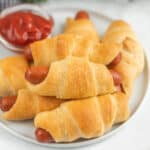 A plate of crescent roll pigs in a blanket—mini sausages wrapped in golden, flaky dough—sits next to a small bowl of ketchup, making this recipe perfect for dipping and sharing.