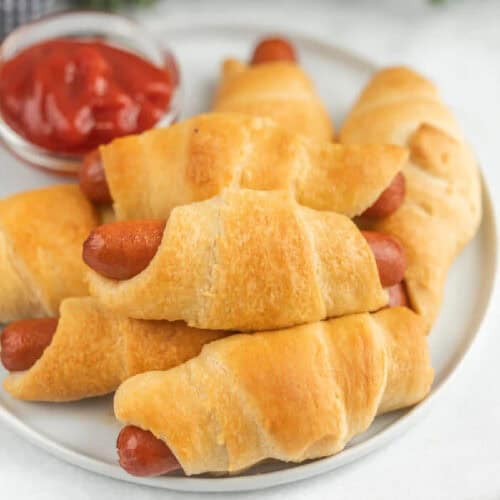 A plate of crescent roll pigs in a blanket—mini sausages wrapped in golden, flaky dough—sits next to a small bowl of ketchup, making this recipe perfect for dipping and sharing.