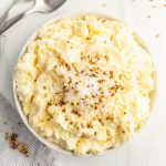 A white bowl filled with creamy, fluffy Pineapple Fluff dessert topped with shredded coconut and chopped nuts; two spoons and a checkered napkin are nearby on a white surface.