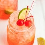 A mason jar filled with a pink, iced Sonic Cherry Limeade, garnished with two maraschino cherries and a lime wedge, with a red-striped straw. Another glass and a lime wedge are visible in the background.