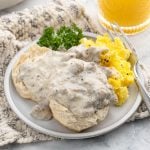 A plate with biscuits and gravy recipe covered in sausage gravy, scrambled eggs topped with black pepper, a parsley garnish, a fork, and a glass of orange juice in the background.