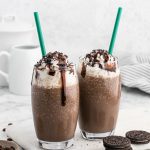 Two Mocha Cookie Crumble Frappuccinos topped with whipped cream, chocolate syrup, and crushed cookies sit side by side in clear glasses with green straws. Oreo cookies and a white pitcher are in the background.