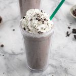 A Starbucks Cookies and Cream Frappuccino in a clear plastic cup is topped with whipped cream, crushed cookie pieces, and a green straw, all set on a marble surface.