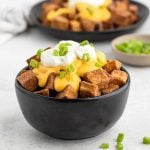 A black bowl filled with crispy diced potatoes topped with melted cheese sauce, sour cream, and chopped green onions just like the Taco Bell Cheesy Fiesta Potatoes Recipe. A blurred bowl of the same dish and small plate of green onions are in the background.