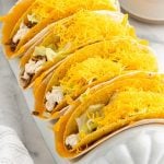 Four hard shell tacos filled with ground beef, shredded lettuce, creamy sauce, and topped with a generous amount of shredded cheddar cheese—reminiscent of the Taco Bell Cheesy Gordita Crunch—arranged in a white taco holder.