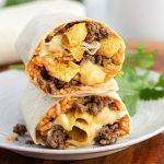 A Taco Bell Frito Burrito cut in half reveals seasoned ground beef, yellow nacho cheese sauce, rice, and crunchy tortilla chips wrapped in a flour tortilla, placed on a white plate with a sprig of cilantro in the background.