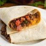 A close-up of a burrito cut in half, revealing a filling of ground beef, melted cheese, diced tomatoes, and green peppers, all wrapped in a flour tortilla on a white plate.