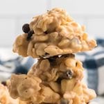 A close-up of two stacked clusters of Avalanche Cookies—white chocolate-covered cereal and raisins—with a blurred background featuring a blue and white checkered cloth.