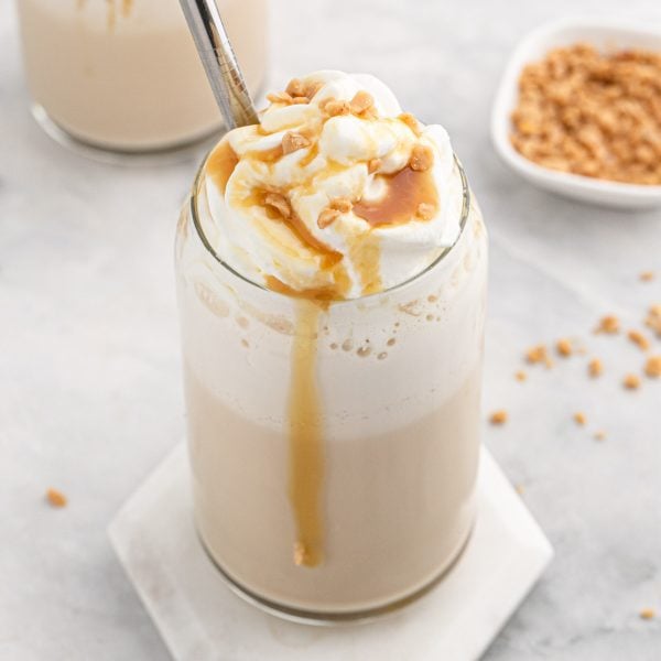 A glass of iced coffee topped with whipped cream, caramel sauce, and toffee bits, just like the Caramel Ribbon Crunch Frappuccino Recipe. Caramel drips down the side, a metal straw stands tall, and a bowl of toffee bits sits in the background.