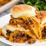 Sloppy Joe Sliders with melted cheddar cheese and seasoned ground beef filling are served on soft sesame seed buns, stacked high on a white plate for the perfect savory bite.