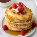A stack of raspberry pancakes topped with butter, fresh raspberries, and syrup sits on a plate, with extra raspberries on the side and a bowl of syrup in the background.
