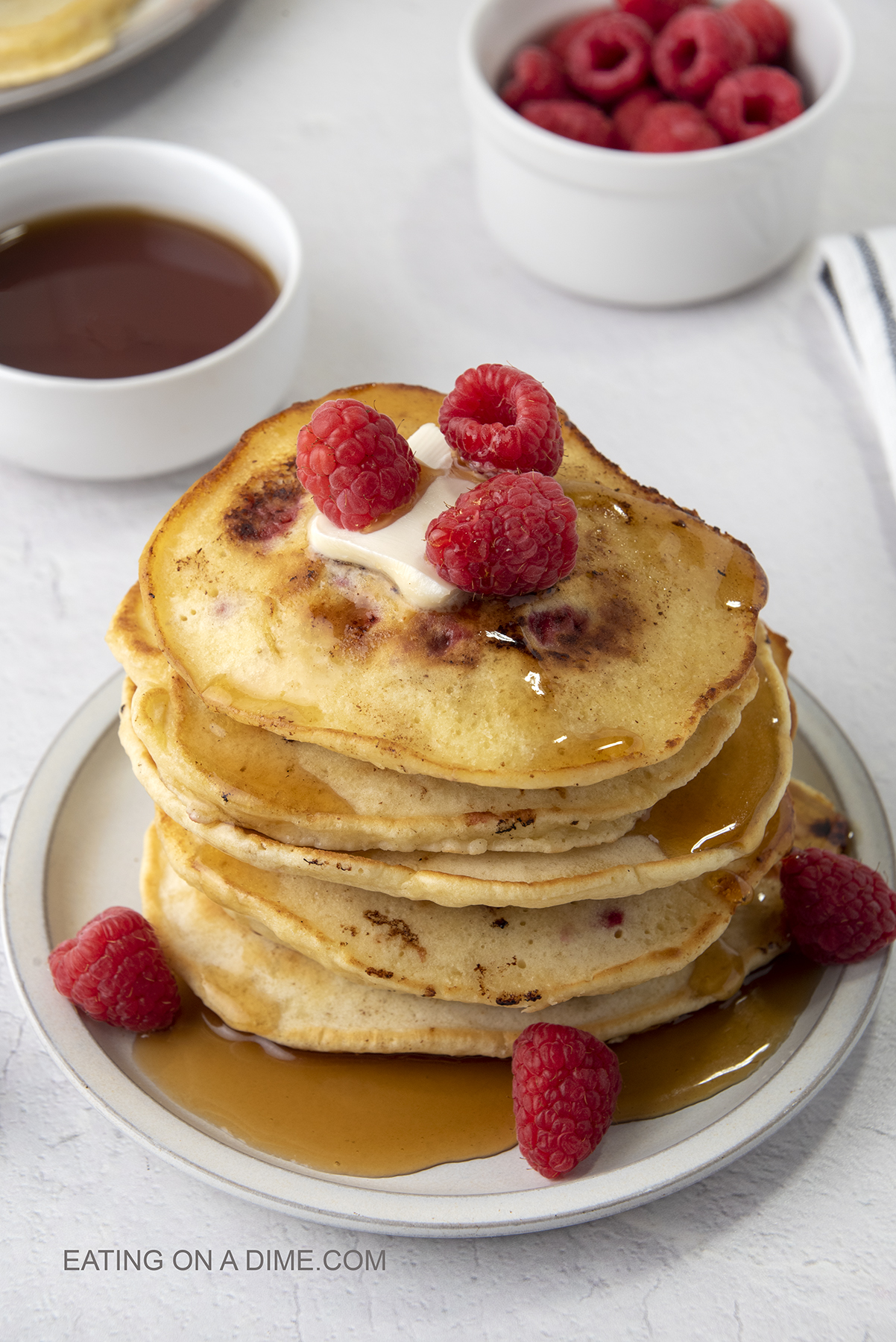Raspberry pancakes stacked on a plate topped with syrup, butter and raspberries