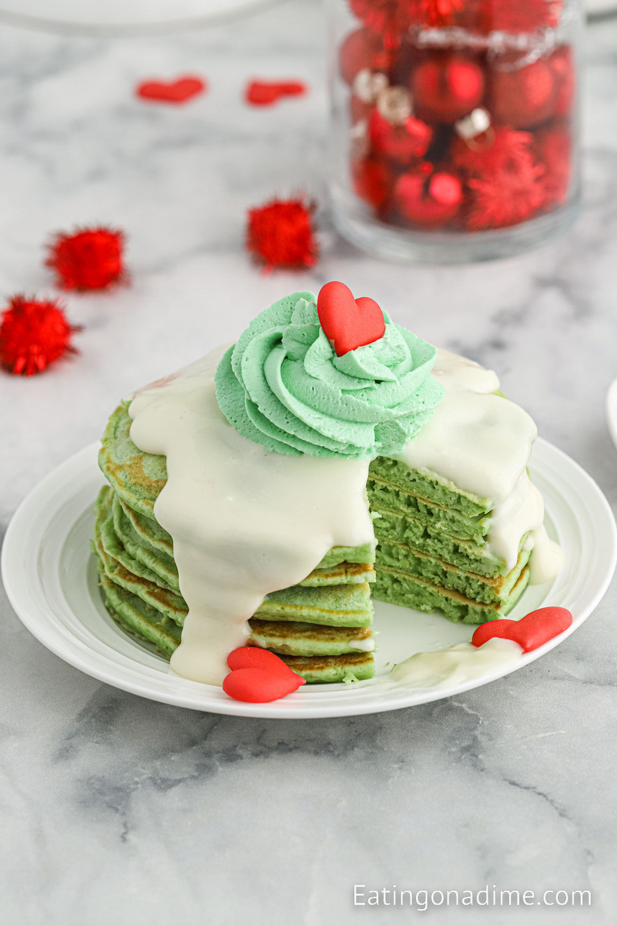 Grinch Pancakes stacked on a white plate topped with cream cheese glaze and green frosting and red heart sprinkles