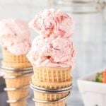 A close-up of a waffle cone with two scoops of creamy strawberry ice cream, with another similar cone blurred in the background. The scene has a bright, fresh, and summery feel.