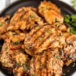 A plate of grilled boneless skinless chicken thighs with visible char marks, garnished with chopped herbs and served on a sleek black dish.