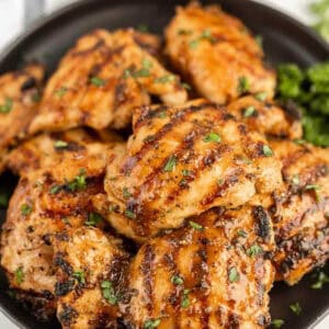 A plate of grilled boneless skinless chicken thighs with visible char marks, garnished with chopped herbs and served on a sleek black dish.