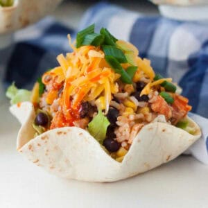 A tortilla bowl filled with rice, black beans, corn, lettuce, and topped with shredded cheese and green onions. The dish is presented on a light-colored surface, with a checkered cloth in the background. This illustrates a Slow Cooker Chicken Burrito Bowl Recipe.