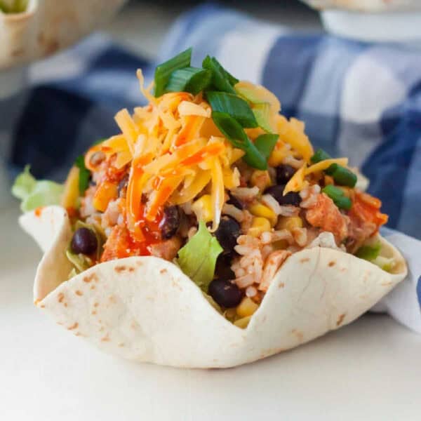 A tortilla bowl filled with rice, black beans, corn, lettuce, and topped with shredded cheese and green onions. The dish is presented on a light-colored surface, with a checkered cloth in the background. This illustrates a Slow Cooker Chicken Burrito Bowl Recipe.