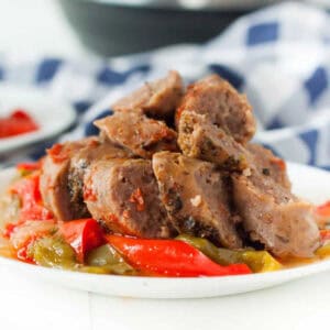 Sliced sausage pieces topped with saucy red bell peppers and green bell peppers on a white plate. The dish is served with a checkered blue and white napkin in the background, suggesting a home-cooked meal context. Instant Pot Sausage and Peppers Recipe.