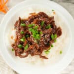 A white plate with fluffy rice topped with Mongolian beef, garnished with chopped green onions and sesame seeds. The dish is rich in color with dark brown sauce covering the beef.