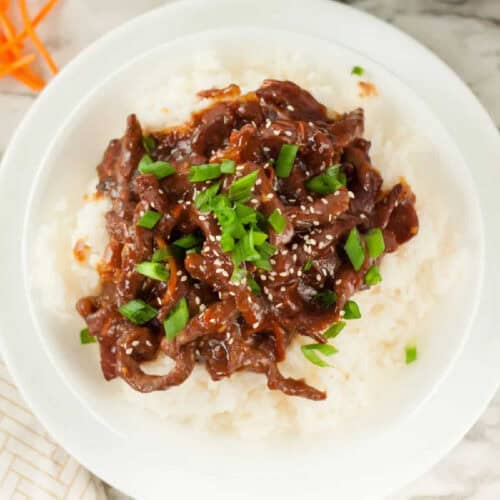 A white plate with fluffy rice topped with Mongolian beef, garnished with chopped green onions and sesame seeds. The dish is rich in color with dark brown sauce covering the beef.