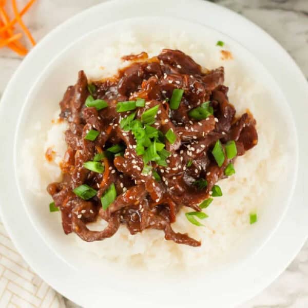 A white plate with fluffy rice topped with Mongolian beef, garnished with chopped green onions and sesame seeds. The dish is rich in color with dark brown sauce covering the beef.