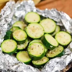 Sliced zucchini seasoned with spices, arranged on aluminum foil. The zucchini slices are round and show a light yellow interior with green skin, ready for grilling. This image supports the recipe for Grilled Zucchini Foil Packs.