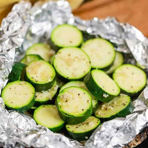 Sliced zucchini seasoned with spices, arranged on aluminum foil. The zucchini slices are round and show a light yellow interior with green skin, ready for grilling. This image supports the recipe for Grilled Zucchini Foil Packs.