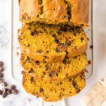 Sliced loaf of Pumpkin Chocolate Chip Bread with chocolate chips on a white surface, surrounded by scattered chocolate chips and a beige-striped cloth.