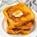 Three slices of pumpkin French toast stacked on a white plate, topped with a pat of butter and being drizzled with syrup. A checkered cloth and silverware are visible in the background.