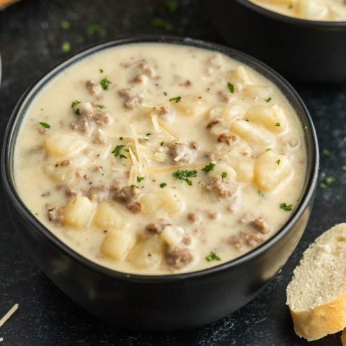 A black bowl filled with creamy sausage potato soup featuring gnocchi, ground sausage, grated cheese, and chopped parsley, served with a slice of bread on the side.