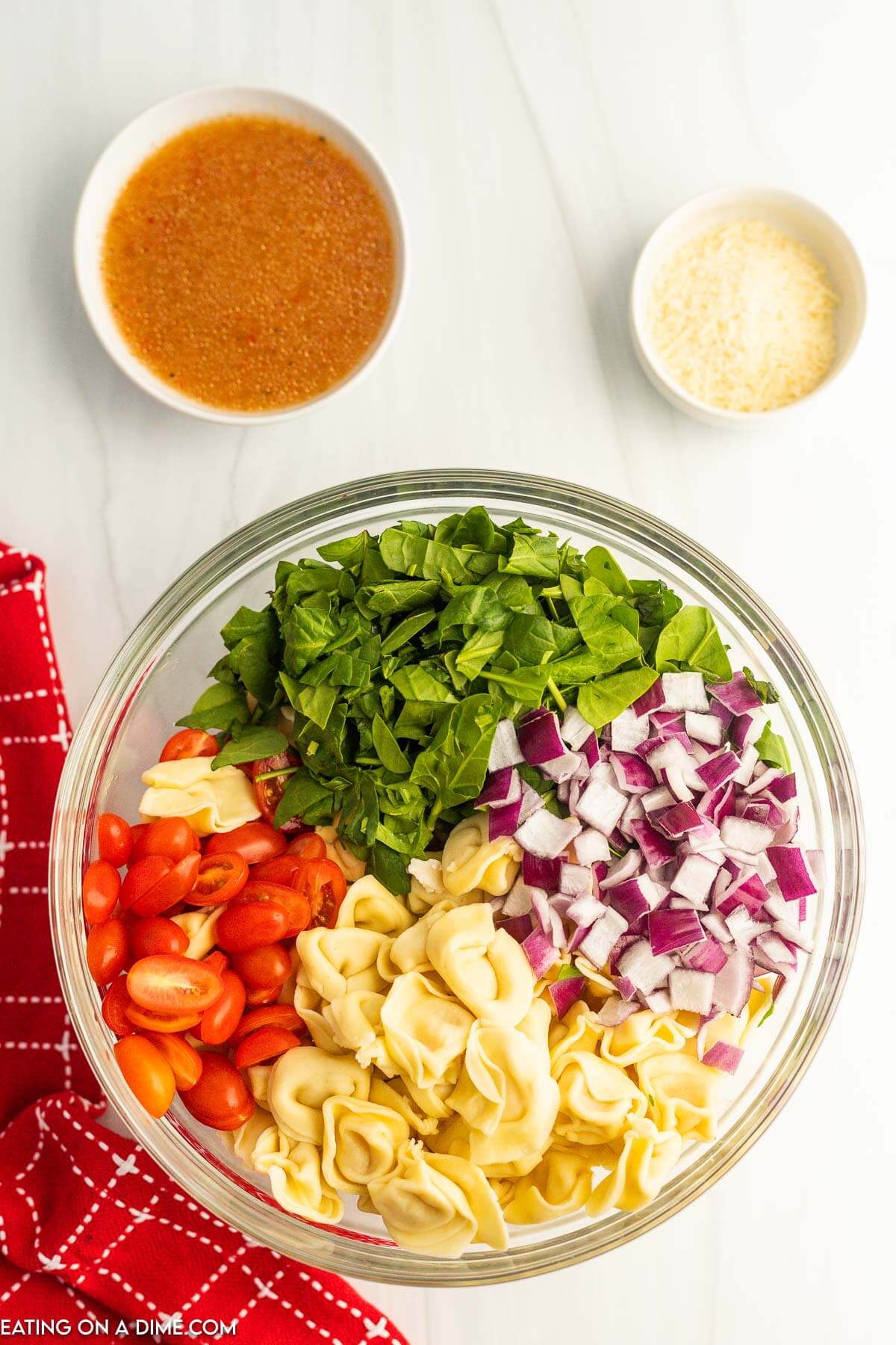 Chopped spinach, slice tomatoes, tortellini, and chopped red onions in a bowl with a bowl of the dressing and cheese on the side