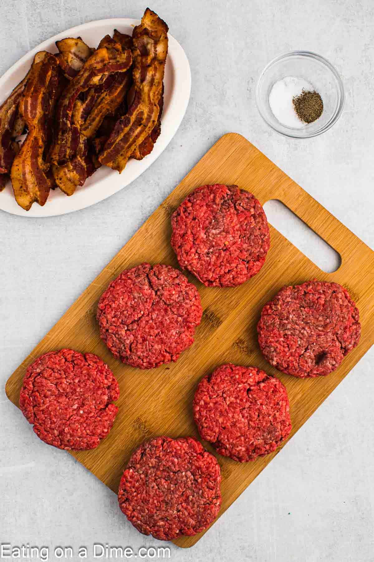 Ground Beef patties on a cutting board with a plate of cooked bacon and a bowl of seasoning