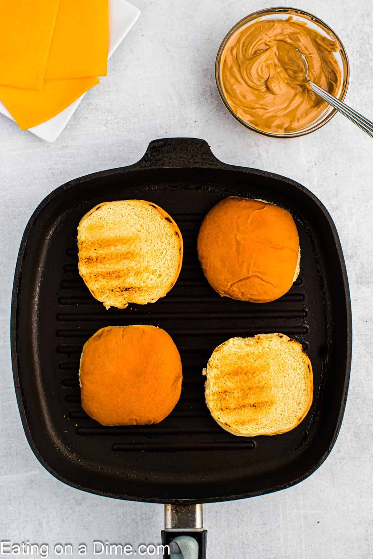 Toasted hamburger buns in a skillet with a plate of slice of cheese and a bowl of a creamy peanut butter