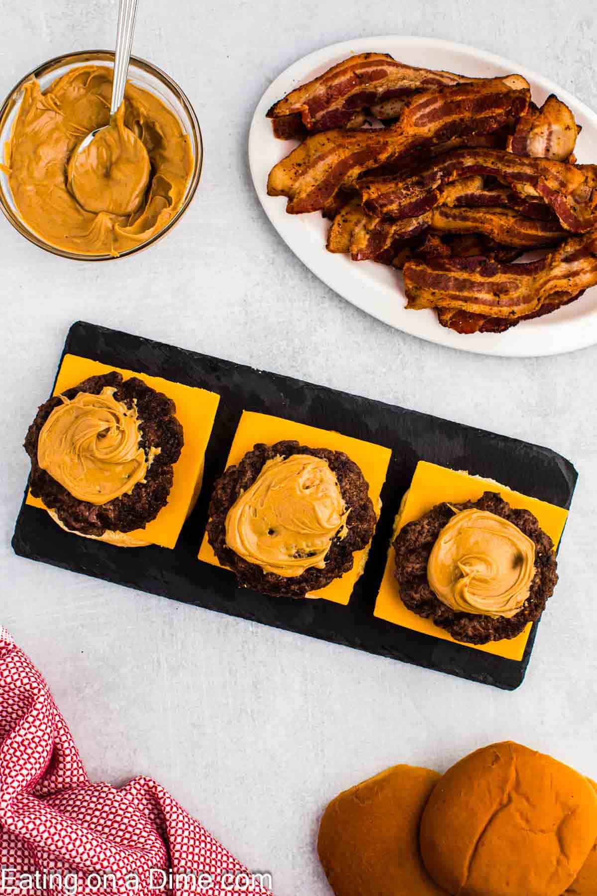 Hamburger patties topped with peanut butter and a plate of bacon and a bowl of creamy peanut butter