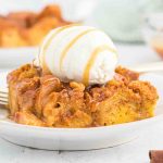 A close-up of Pumpkin Bread Pudding drizzled with caramel sauce, topped with a scoop of vanilla ice cream, on a white plate.