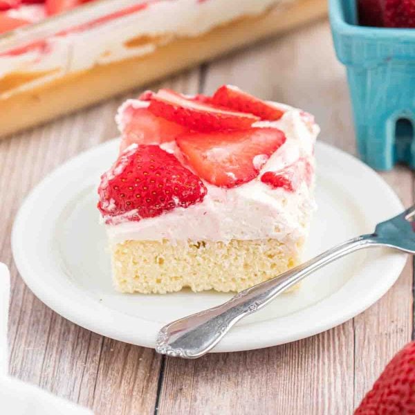 A slice of Strawberry Shortcake Bars with a light yellow base, creamy white frosting, and fresh strawberry slices sits on a white plate with a fork. In the background, thereโs a tray and a blue container.