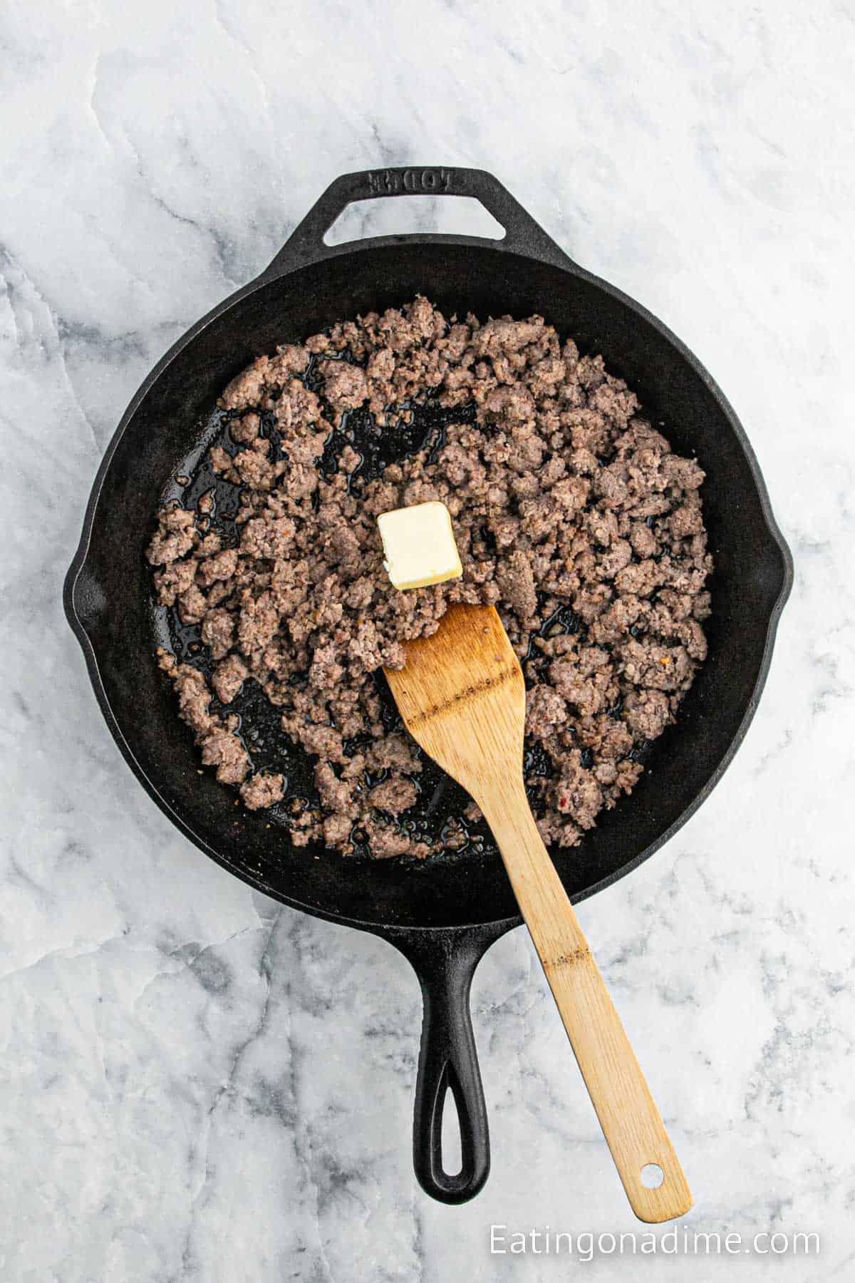 Cooked ground sausage in a cast iron skillet with a slice of butter on top with a wooden spoon