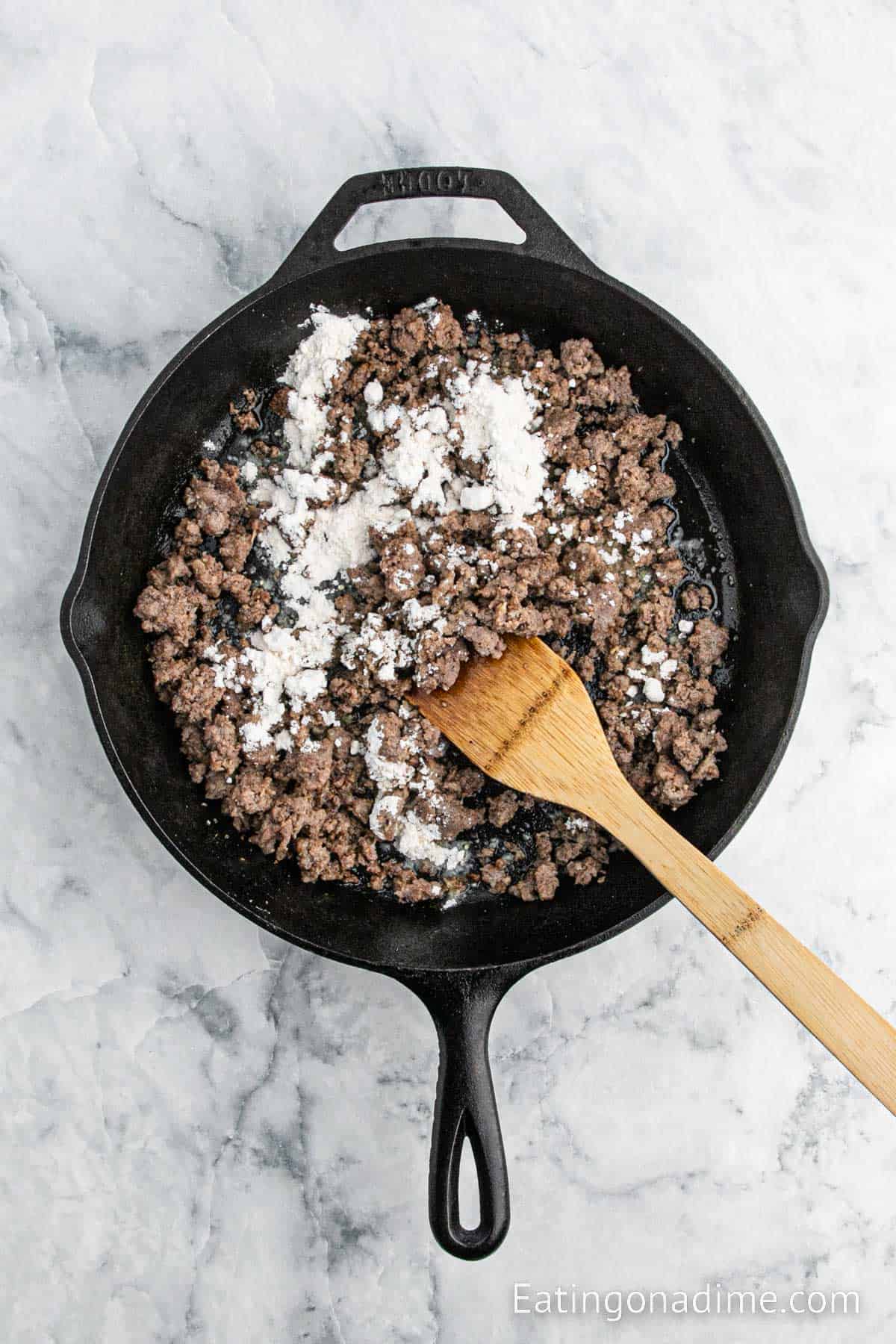 Flour sprinkled on top of the cooked sausage in the cast iron skillet with a wooden spoon