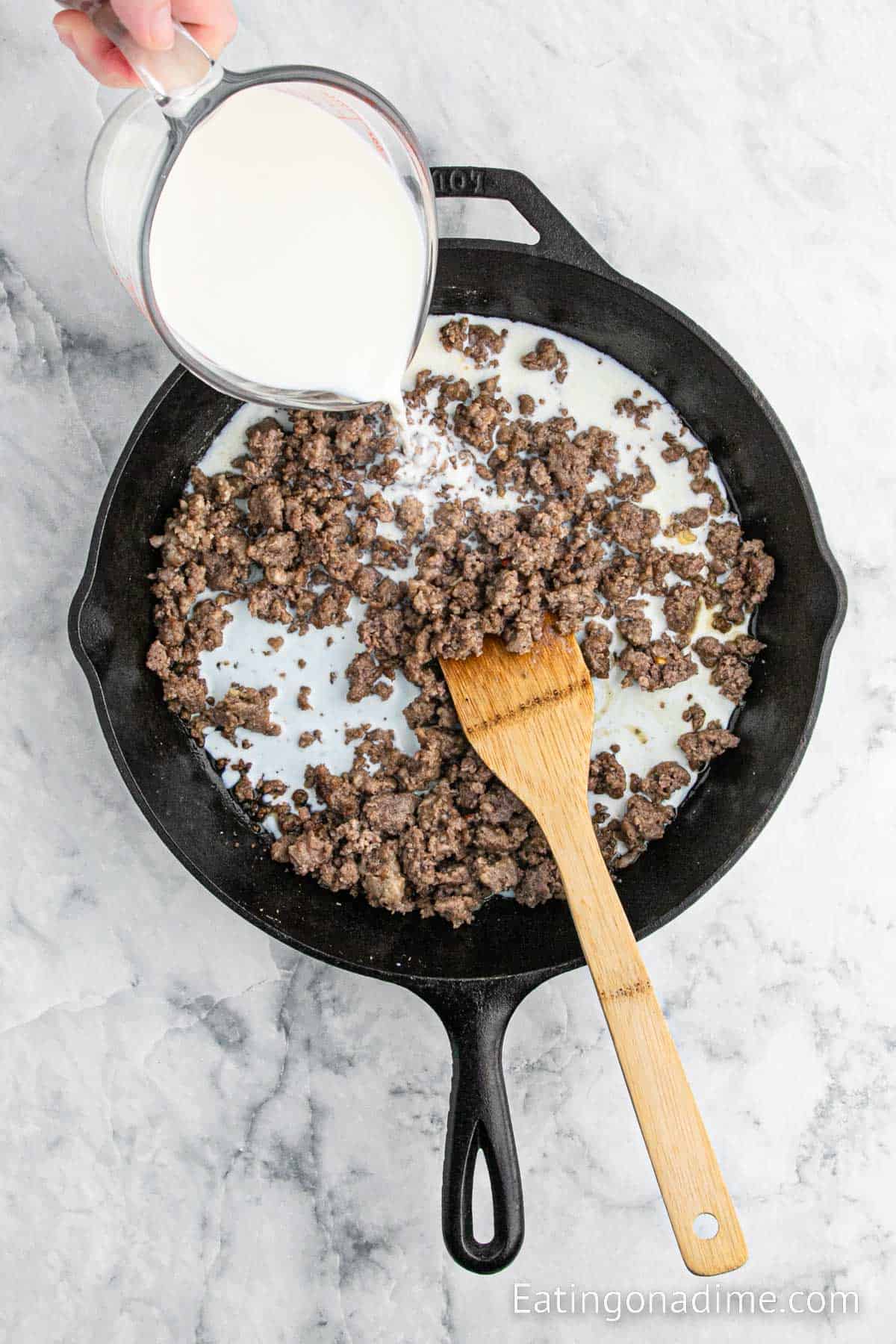 Pouring milk into the cast iron skillet with ground sausage and a wooden spoon