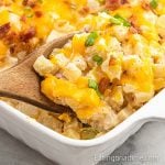 A wooden spoon scoops this loaded baked potato casserole, featuring cheesy hashbrown casserole topped with melted cheese, bacon bits, and green onions from a white baking dish.