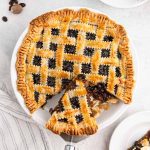 A baked mincemeat pie recipe features a golden, crimped lattice crust filled with dark fruit filling. One slice is being lifted out, revealing the juicy interior. The pie sits on a light surface with a striped towel nearby.