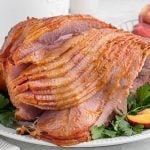 A sliced, golden brown Peach Glazed Ham Recipe is served on a white plate, garnished with fresh parsley and juicy peach slices. The ham glistens with a shiny, caramelized glaze.