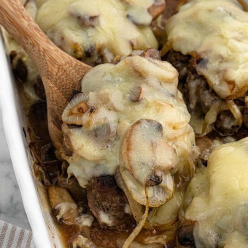 A close-up of Texas Roadhouse Roadkill—baked hamburger steaks topped with melted cheese, sautéed mushrooms, and onions in a white baking dish. A wooden spoon is lifting one of the juicy steaks.