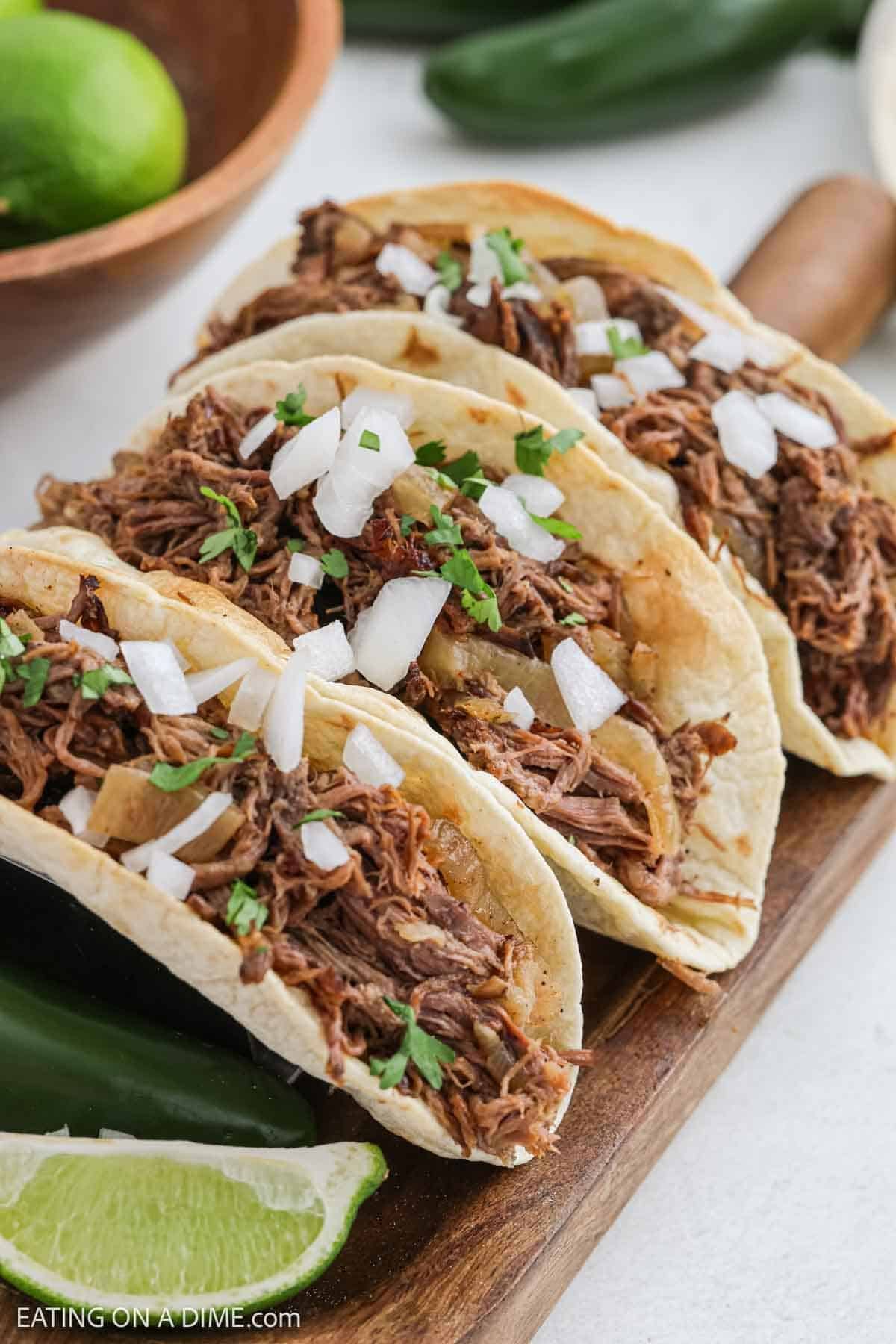 Three shredded beef tacos made with a slow cooker beef carnitas recipe are topped with chopped onions and cilantro, arranged on a wooden board with lime wedges and a green pepper on the side.